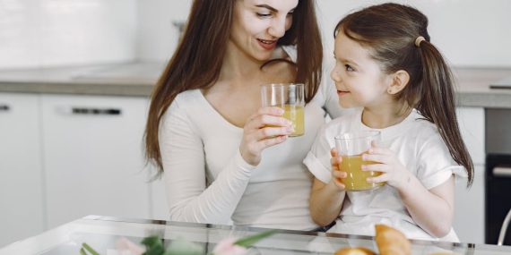 Happy mother and daughter in casual clothes drinking juice and looking at each other while having breakfast together in contemporary kitchen